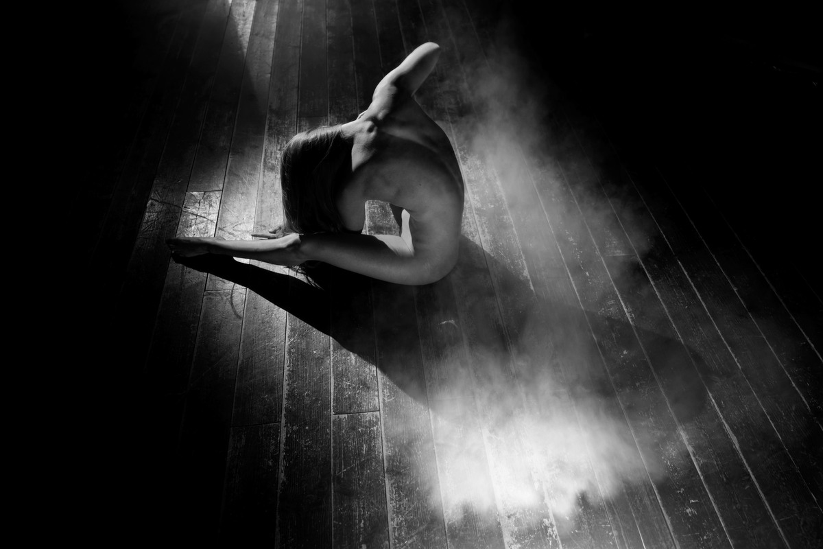 Nude figure seated on a wooden floor amid a pale cloud of smoke.