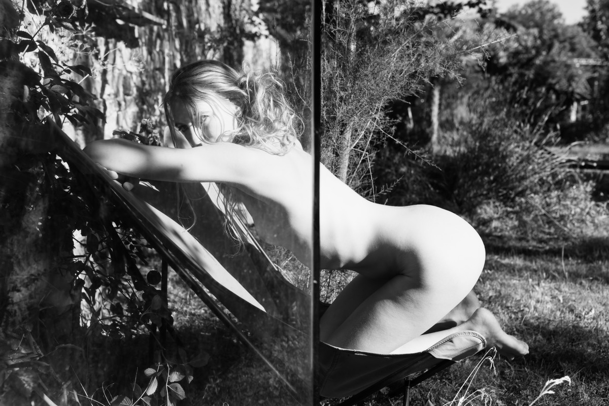 Nude figure bending across a fragmented reflective surface outdoors.