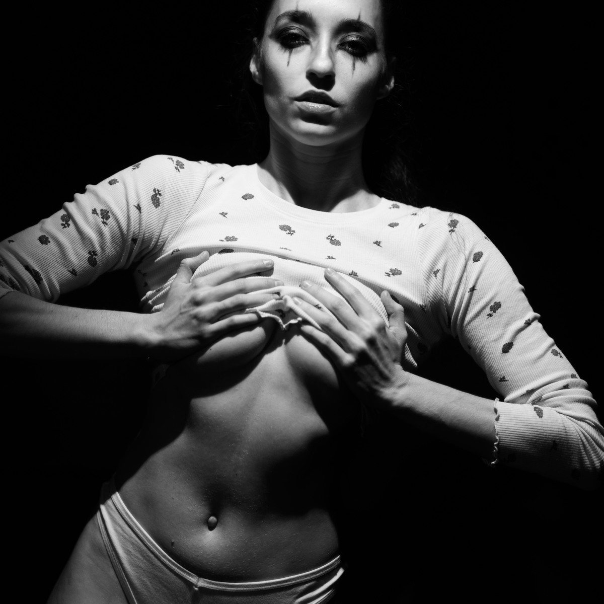 Woman lifting a patterned top to reveal her midriff against a black background.