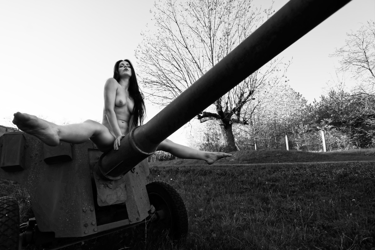Nude woman sitting on a large cannon barrel outdoors.