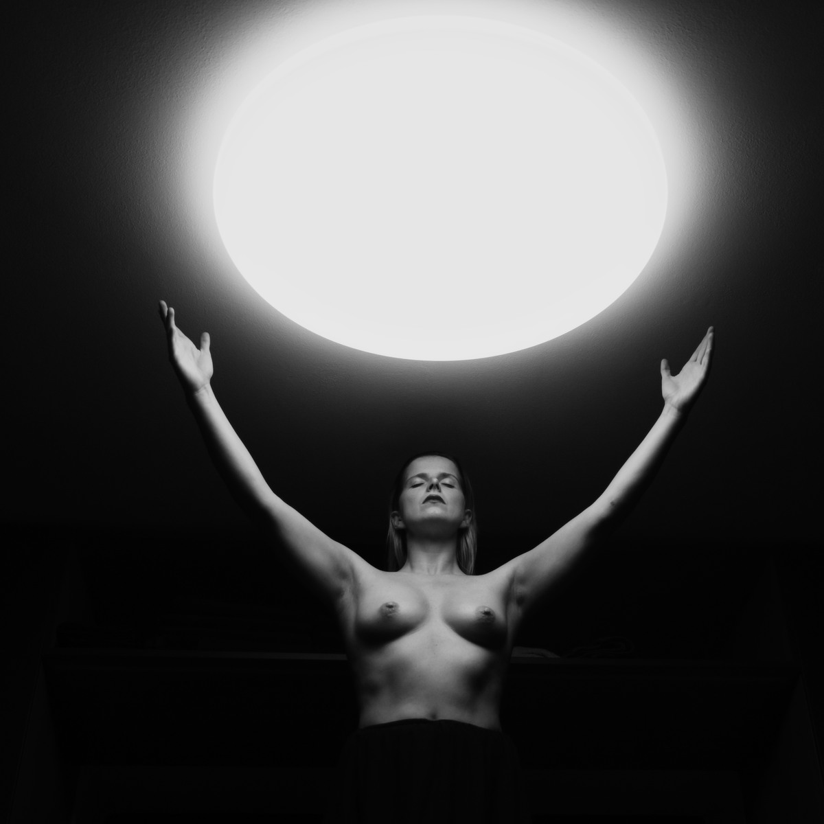 Nude figure with raised arms beneath a large circular ceiling light.