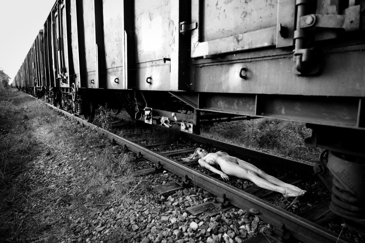 Nude figure lying alongside railway tracks beneath freight cars.