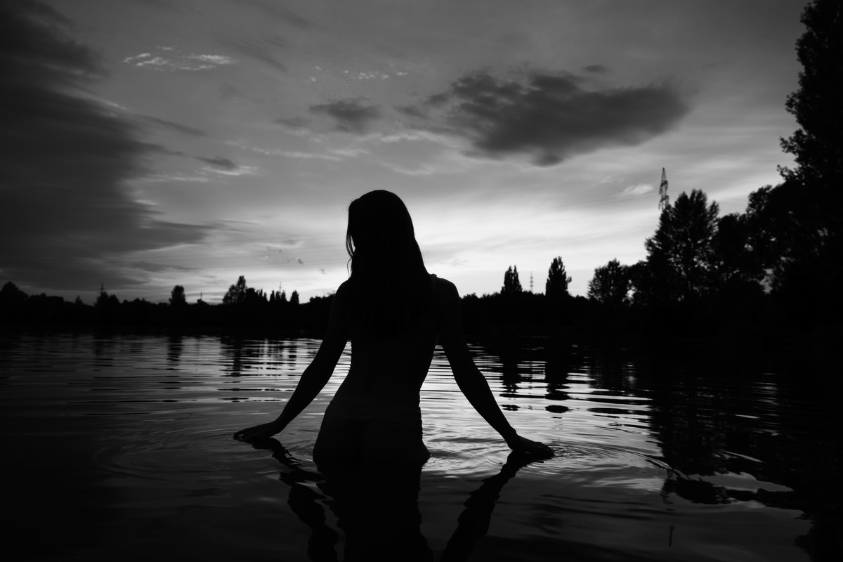 Silhouetted nude figure standing waist-deep in water at dusk.